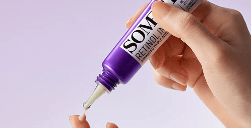 دست یک فرد در حال استفاده از کرم دور چشم بنفش SOMEBYMI با اپلیکاتور فلزی، در پس‌زمینه‌ای به رنگ بنفش ملایم.